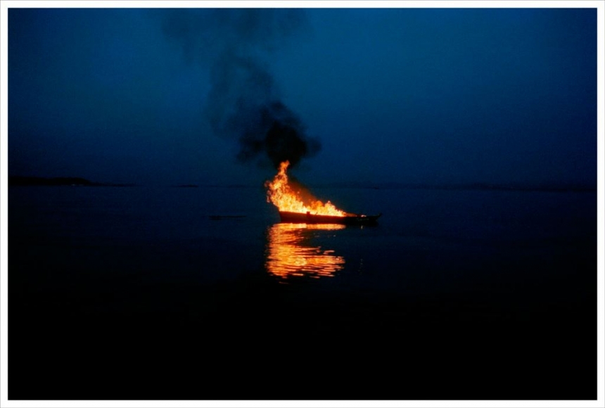a photograph from video art by thiago rocha pitta showing a boat on fire in the water