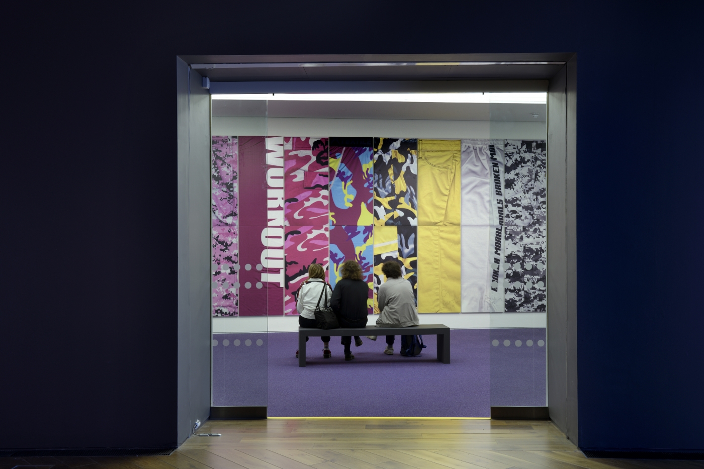 Three people sit on a black bench with their backs turned in front of a multicolored, graphic wall piece.
