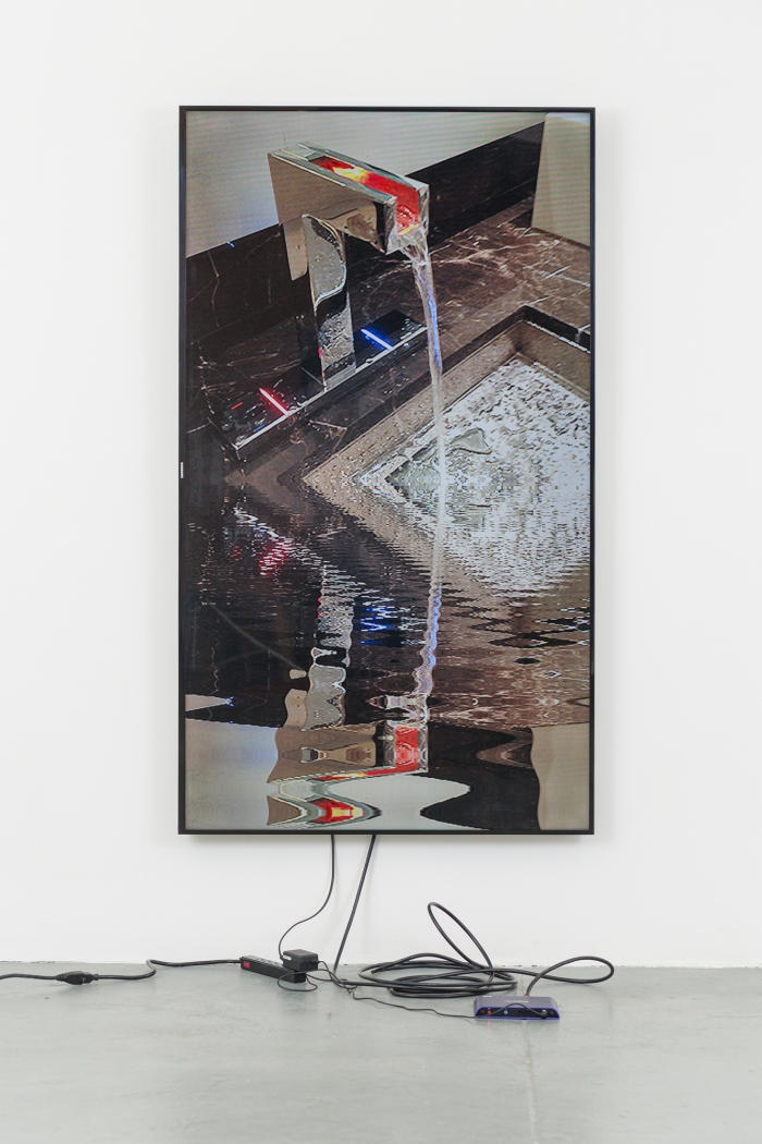 A vertically aligned television monitor displaying a running sink mounted on a granite countertop. The water reflects the image of the sink, distorted.
