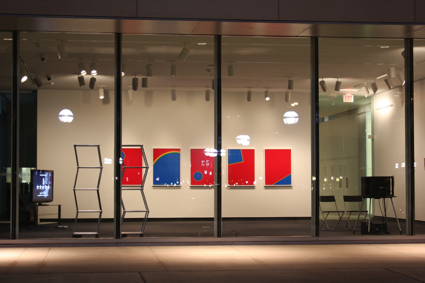 Through four paneled windows, 5 red and blue canvases with geometric shapes are visible, along with two vertical silver trolleys constructed at disagonals, and two monitors on each side of the room.