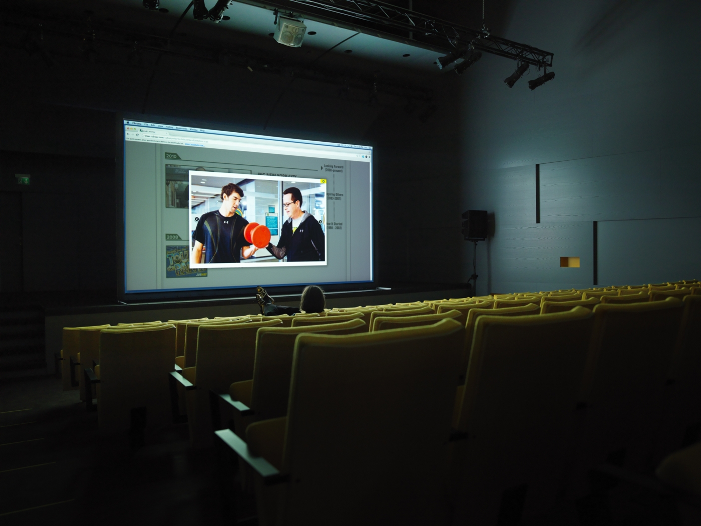 An empty theater with yellow chairs projecting a Macbook screen, displaying a stock image of two men in a gym, one holding a bright red dumbbell.