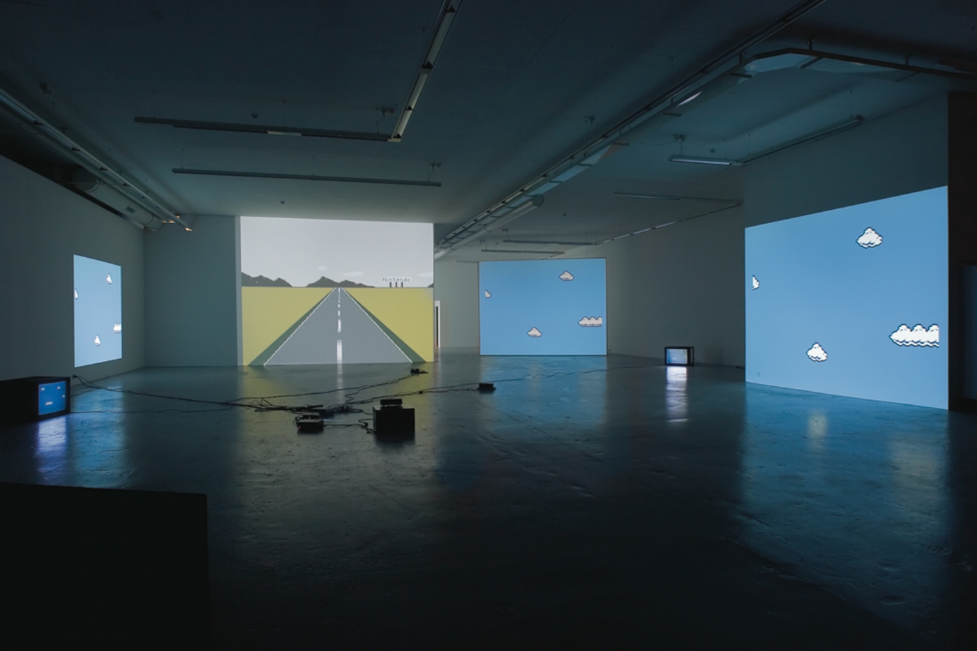 A multiple channel projection on four walls. From left to right: pixelated clouds in against blue background, an empty pixelated street perspective, two projections of pixelated clouds against blue backgrounds. Several analog monitors are placed on the floor next to three of the projections.