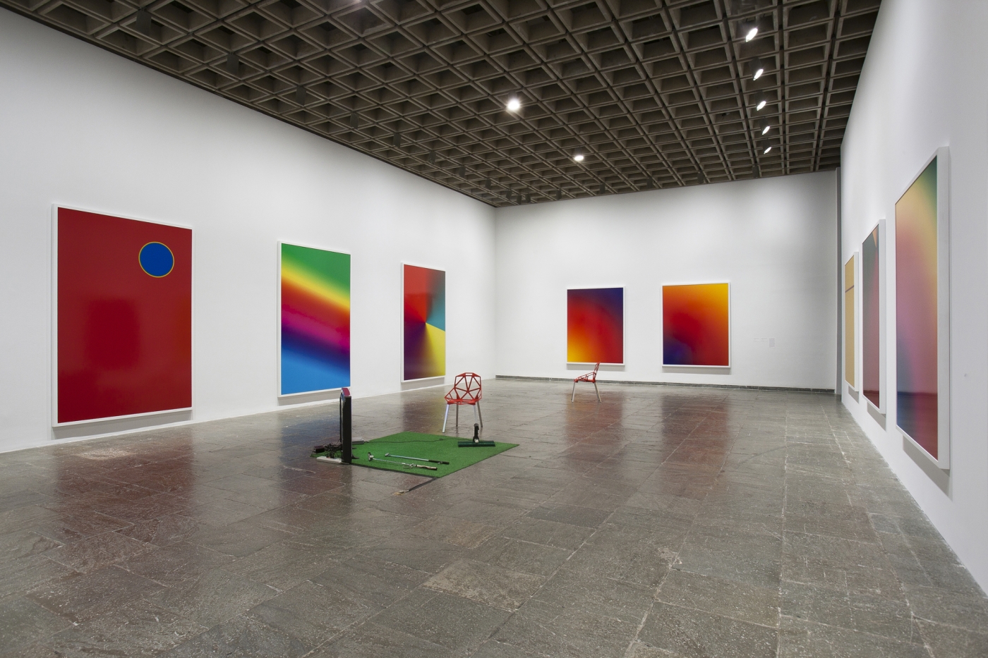 Eight multicolored gradient panels along the surface of three walls. In the center of the room is a small green turf with a geometric chair and assorted small objects.