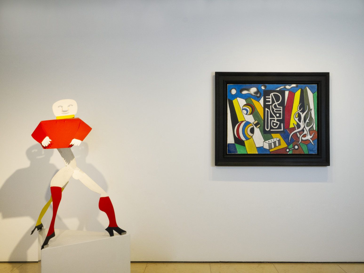 Installation view of Masterworks. This image shows a sculpture by American sculptor Alexander Calder part of the Critter series, titled "Haute Couture" and produced in 1976. It depicts a man standing on three legs. The top part of the sculpture, including the bust and head was produced as an independent piece that is simply seated on the top and revolving when it catches the wind. On the right is a paiinting by fernand Leger, representing in a palette of primary colors, geometric forms and objects, in a abstract way, depicting an imaginary space. The painting is titled "Objets dans l'espace" and was produced in 1931, the year he discovered NY city for the first time and manifests his impression the city had on him.