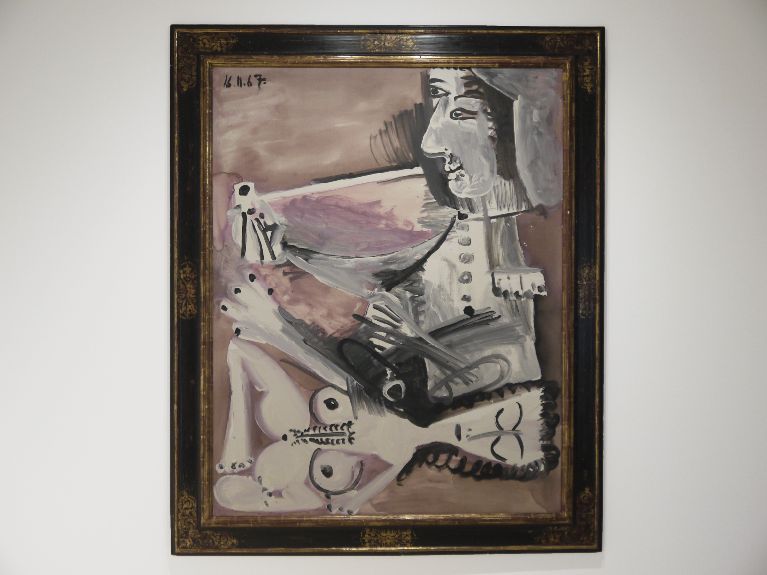 This photo features a painting by Pablo Picasso which title is "Homme à la pipe et nu couché", 1967. It represents a couple, a man seated smoking a pipe and a naked woman laying down by his side. Both characters fully occupy the canvas. The overall palette color is mauve, the characters are depicted in grey and white tons and their features, hair and attire are made of large black lines.