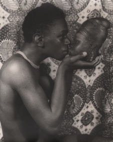 Waist-up portrait of the figure, shirtless, in profile, holding a traditional mask to face him so they are nose-to-nose.