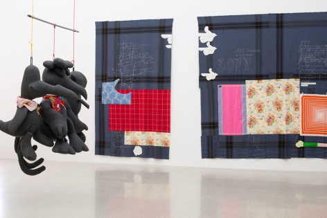 Installation view of von Bonin's show at MUMOK featuring three large hanging stuffed birds, each wearing colorful scarves. Two patch-work cloth "paintings" hang in the background.