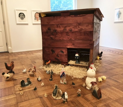 Installation work by February James consisting of a wooden chicken coop with faces painted on each side and tens of found ceramic chickens on the floor.