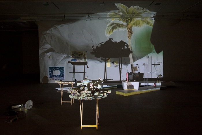 Bag's Circle, 2012, Single-channel video installation with four acrylic-on-canvas paintings, painted foam sculptures, CD rack, plastic cups, sticks, plexiglass, tape, clay, three folding tables, sequined throw, extendable fork, Continuous loop, Dimensions variable