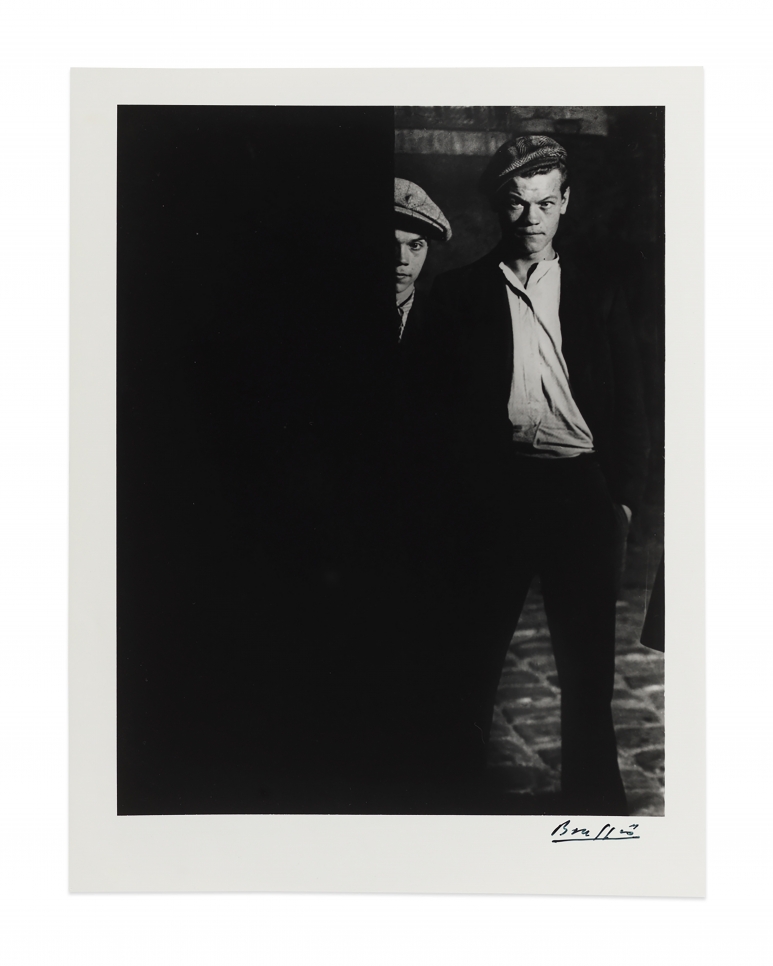 Black and white photographic by Brassaï featuring two men wearing hats. Half of one man's face is showing behind a barrier, and the other's full face is showing