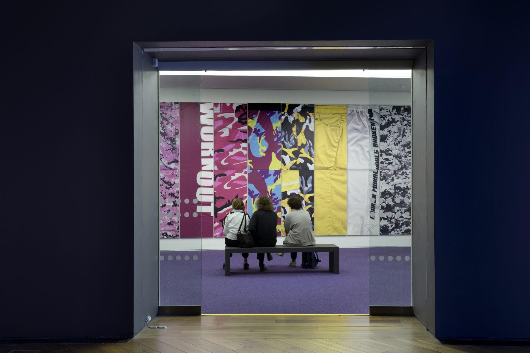 Three people sit on a black bench with their backs turned in front of a multicolored, graphic wall piece.