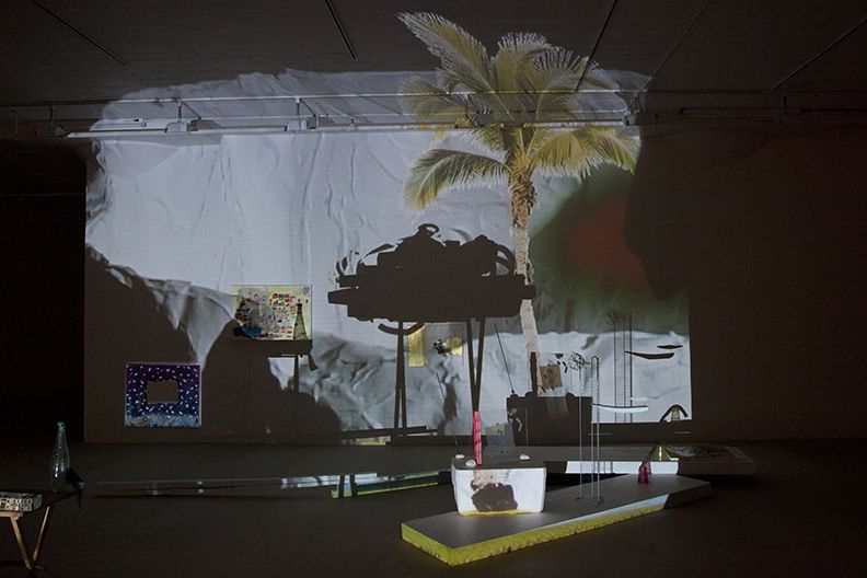 Bag's Circle, 2012, Single-channel video installation with four acrylic-on-canvas paintings, painted foam sculptures, CD rack, plastic cups, sticks, plexiglass, tape, clay, three folding tables, sequined throw, extendable fork, Continuous loop, Dimensions variable