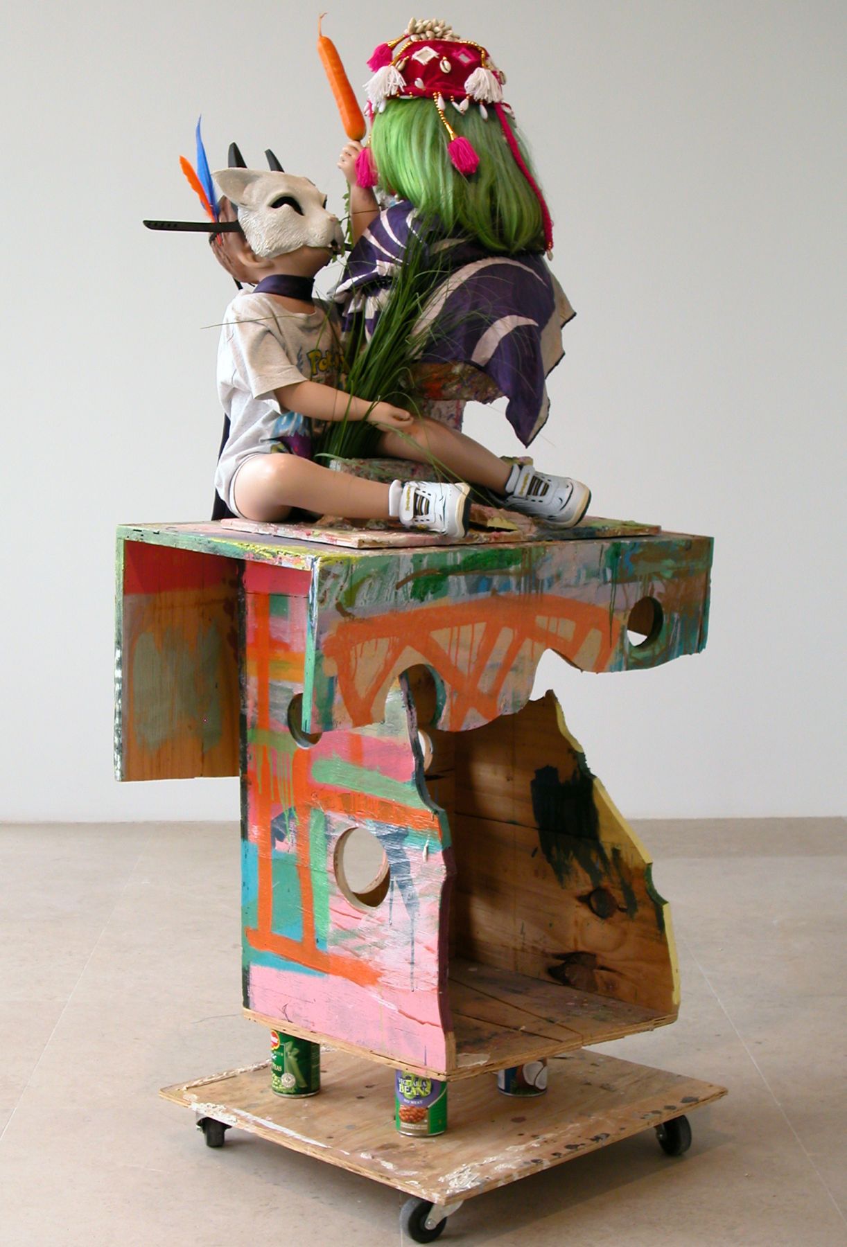 Rachel Harrison I'm with Stupid, 2007 Wood, polystyrene, cement, acrylic, child mannequin, papier-mâche skull, green wig, festive hat, Spongebob Squarepants sneakers, Pokemon t-shirt, wheels, canned fruits and vegetables, fake carrot, fake feathers, fake grass, Batman mask, cat mask, necktie, scarf, and plastic beads 65 x 31 x 24 inches 165.1 x 78.7 x 61 cm