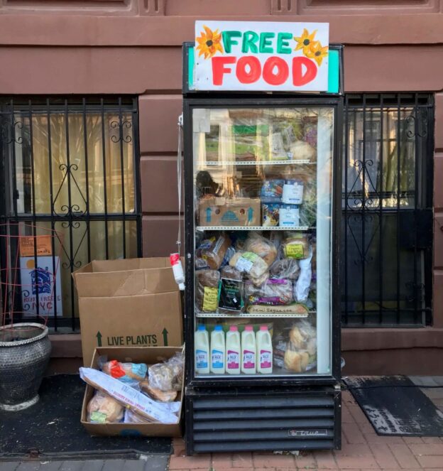 Photograph of a community fridge full o ffresh food. On top of the fridge, a sign recites "Free Food" in colorful letters