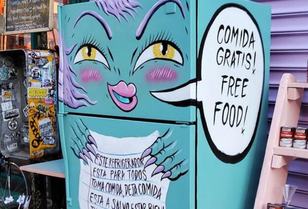 Photo of a community fridge. The fridge is painted light blue and decorated to look like a smiling monster. A balloon from the mouth of the monster recites "Comida gratis! Free Food!"