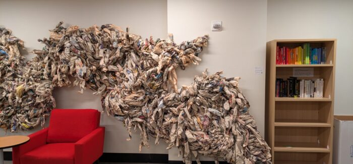 Image of newspaper-based artwork hanging on the wall in the background with a red chair and bookshelf in the foreground.