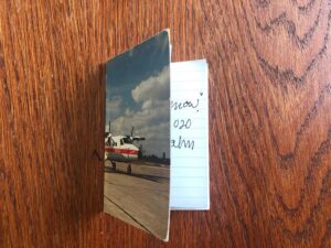 A postcard showing a helicopter on a runway with blue skies is used as a cover wrapping a handwritten journal. In this image, the notebook is closed, so only half of the postcard shows. A glimpse of the interior pages is visible.