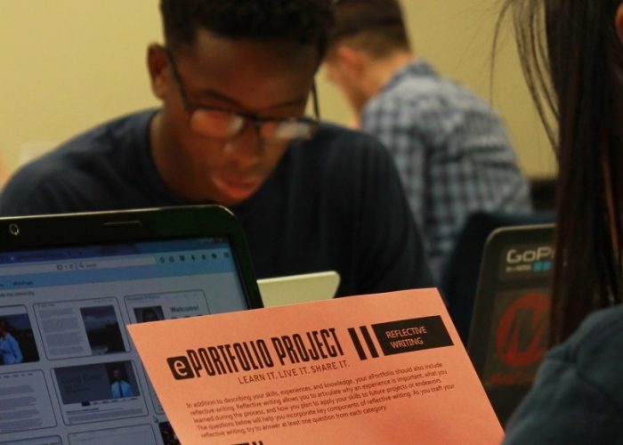 The image features a person, viewed from behind on the right side, reading a paper titled "ePortfolio Project: Reflective Writing." Behind the paper, a laptop is open. Several other students work in the background.