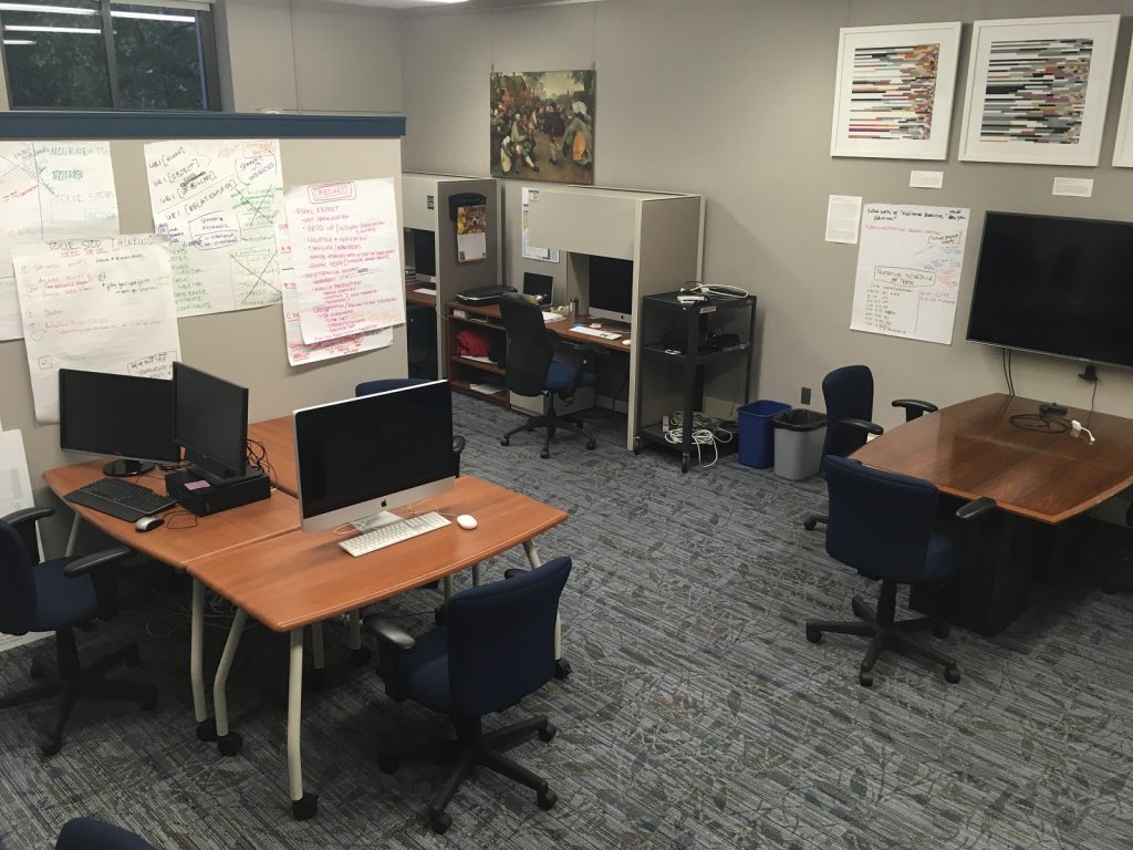 A photograph of the interior of the Visual Media Workshop at the University of Pittsburgh, taken in October 2017, that shows a grouping of desks with computers, but that is not currently populated with people.