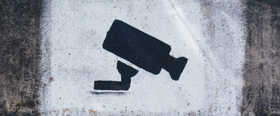 A black security camera stencilled onto a painted white cement background that is rectangular in shape. Around the white rectangle is more cement that is more dirty and grimy.