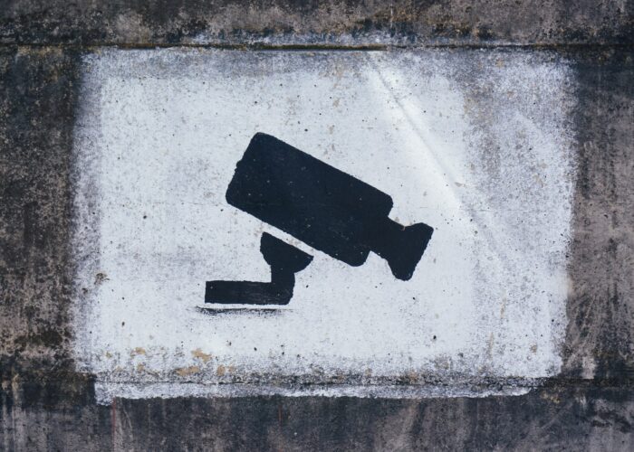 A black security camera stencilled onto a painted white cement background that is rectangular in shape. Around the white rectangle is more cement that is more dirty and grimy.