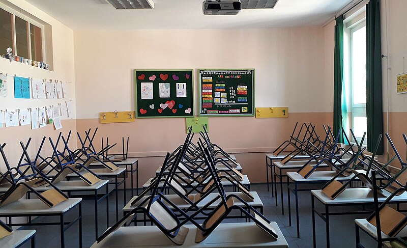 An empty classroom with children's drawings on the walls and chairs stacked atop rows of desks