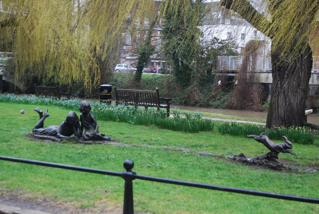 green lawn with a willow tree featuring a statue of a young girl and and older girl with a book to the right is a running rabbit statue