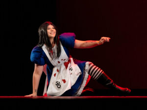 A dark haired woman leans on the ground with one hand and leg in a black and white tights, boots a blue puffed dress sleeve dress with a bloody white apron over it and a pocket with a black symbol.