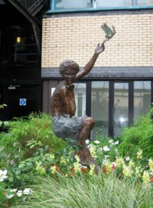 Statue of a boy with arm upraised to a small fairy, building and flowers in background