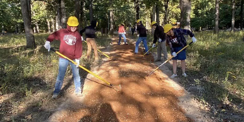 Six individuals taking part in trail maintenance in a forest, all with rakes in their hands spreading soil.