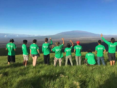 A group of ten all in green shirt stand with their back facing the camera looking off into the mountains.