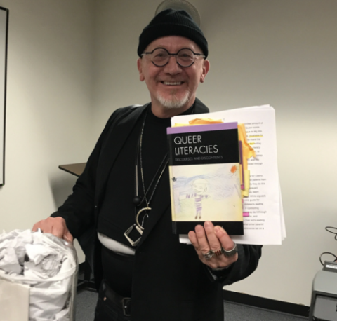 Professor Mark McBeth, wearing glasses, a black beanie, and dark clothing, smiles warmly while holding his book Queer Literacies: Discourses & Discontents.