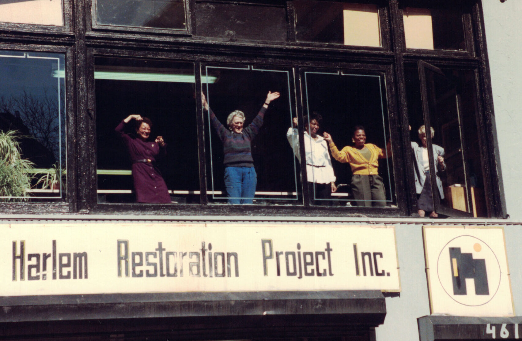 A color photo of a small group of people waving from behind a second-story window. The sign on the large building reads, "Harlem Restoration Project, Inc.: