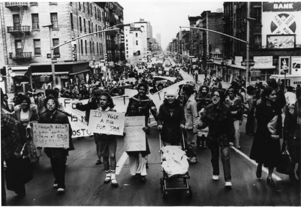 A black and white photograph of a diverse group of young people holding signs, including a woman pushing a stroller, marching in the streets of Manhattan.