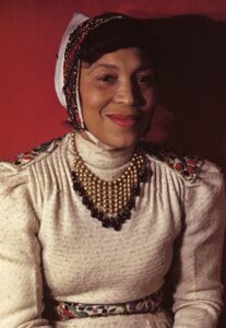 A color photograph of anthropologist Zora Neale Hurston, a smiling Black woman wearing a white dress with jeweled embellishments. Her bright red lipstick matches the red wall behind her.