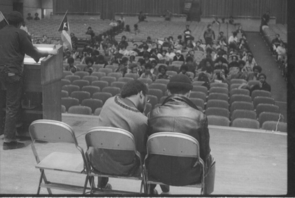 A black and white photograph shows the backs of two people sitting on a stage facing a partially full auditorium.