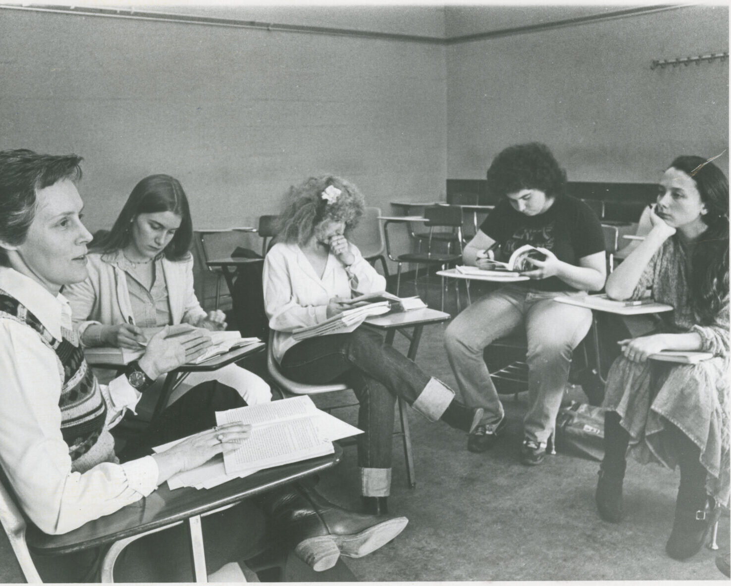 A black and white photo depicts a group of five women wearing vintage, 70s-era clothing, sitting in a circle in a classroom. 
