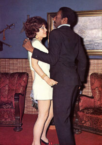 A black man and a white woman stand in front of a painting. They are smiling and laughing.