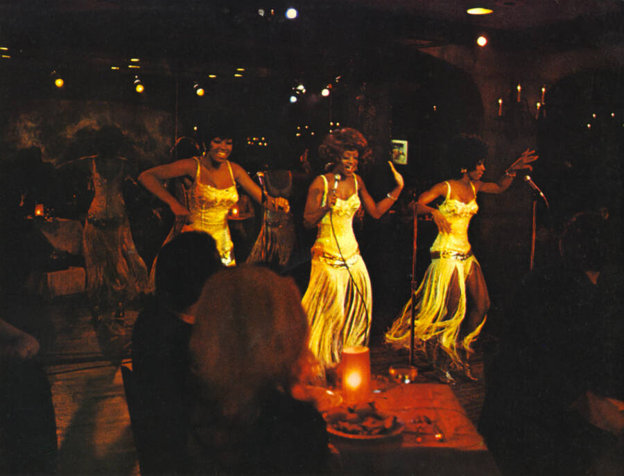 Three women of color perform on a stage at a discotheque.