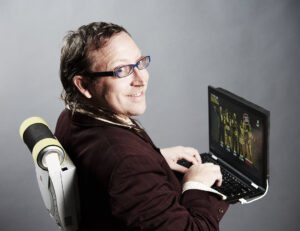 Nick Normal smiles over his shoulder while typing at a laptop that appears to display a computer game.