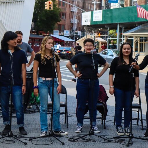 Photo of 6 people wearing jeans and black t-shirts, standing in front of microphones on the sidewalk of a city street