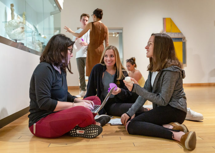 Students sitting on the ground at a museum smile and discuss the 3D printed antiquity replicas they're discussing.