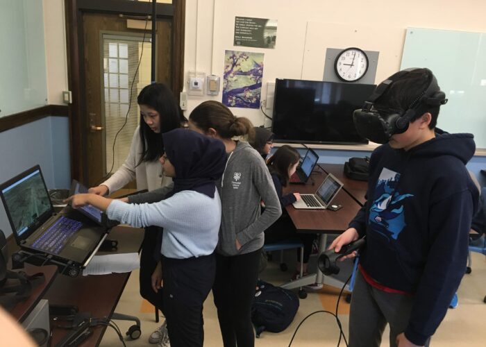 Three students stand at the front of a classroom. They are pointing at a computer and consult a map. A student stands behind them wearing virtual reality equipment.