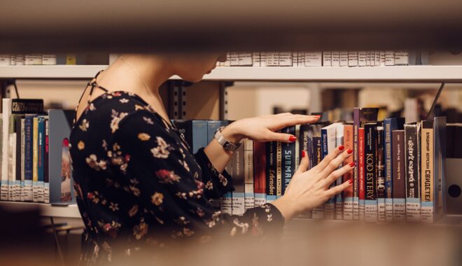The view peering through the stacks sees a person, face obscured, browsing the books.