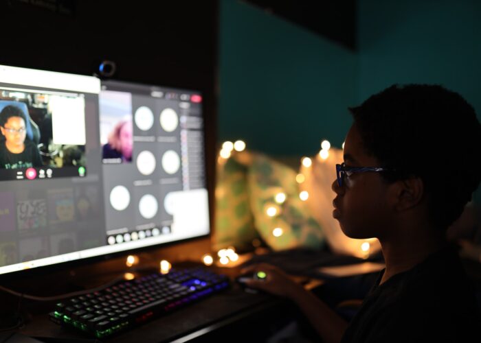 A young person, in shadow and profile, gazes at their own video feed as they take a class online from home, faced with a webcam.
