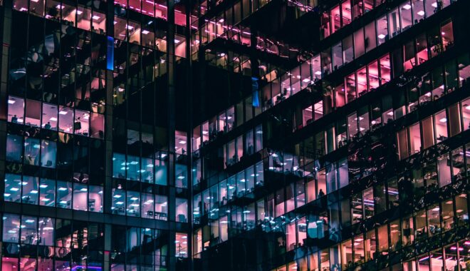 The glass windows of two intersecting wings of an office building are lit up at night.