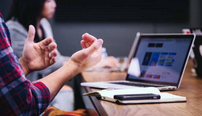 Two people discuss by a computer and smartphone, gesticulating with their hands.
