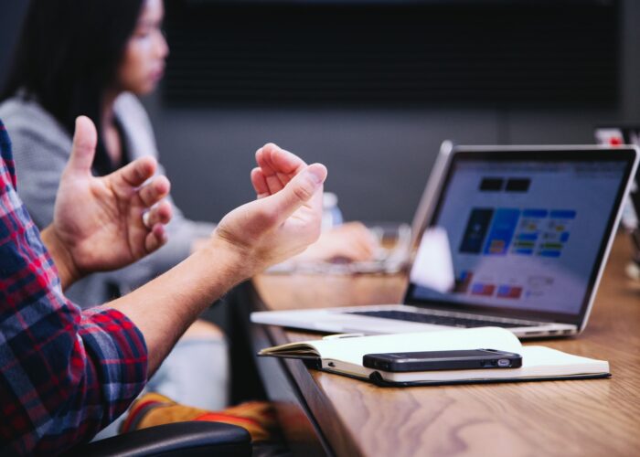 Two people discuss by a computer and smartphone, gesticulating with their hands.