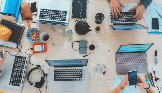 A variety of laptops sit, viewed from above, on a table, worked by hands also using external hard drives, phones, and headphones, alongside snacks and tea.