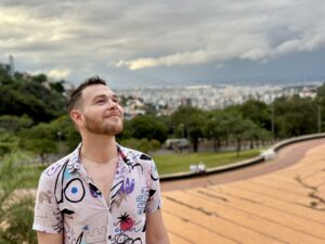 Image of a person standing in the left side of the frame wearing a collared shirt with their head angled up towards the sky with green space directly behind them and a city just behind the trees in the far background.